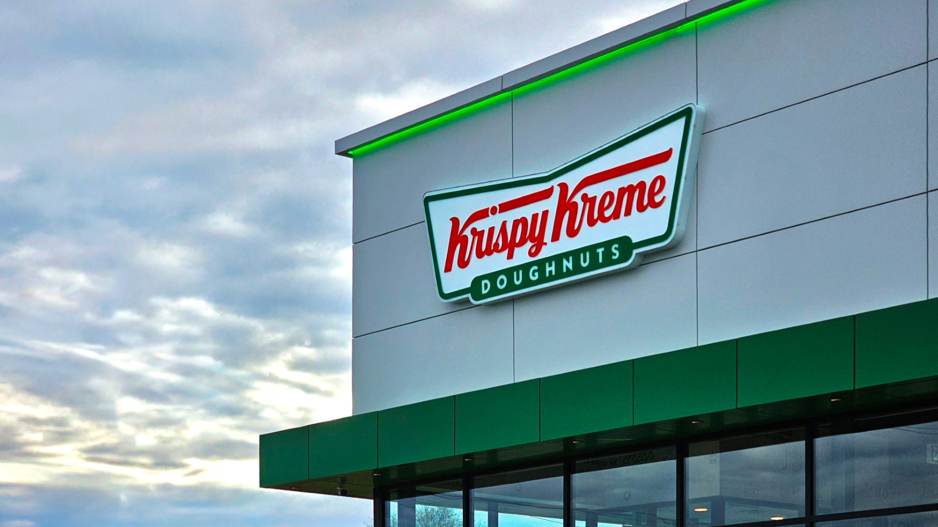 A Krispy Kreme Doughnuts storefront with a light gray facade, green trim, and large glass windows. The illuminated Krispy Kreme logo is mounted high on the building beneath a cloudy sky.